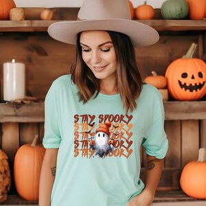 May include: A light teal t-shirt with the words "STAY SPOOKY" in orange and white. The shirt features a cartoon ghost wearing a hat and holding a pumpkin. The background includes pumpkins and a wooden shelf.