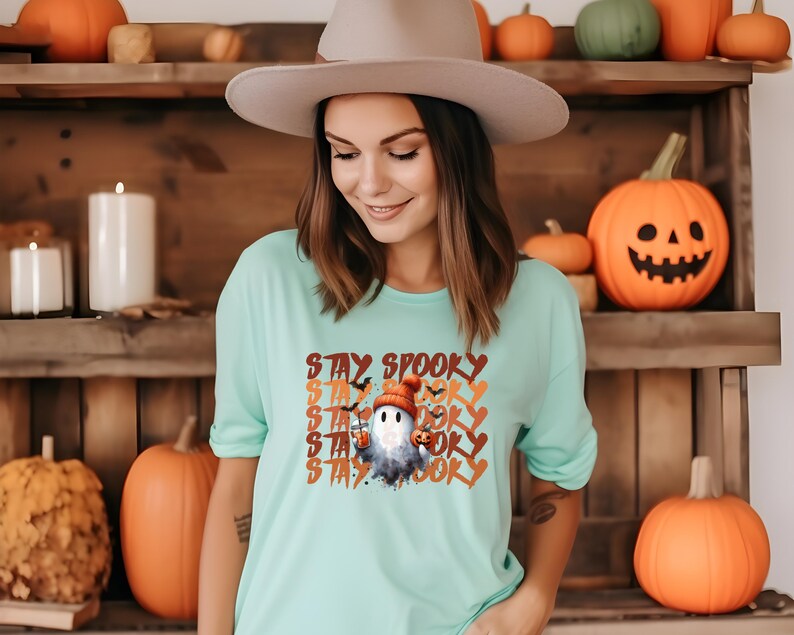 May include: A light teal t-shirt with the words "STAY SPOOKY" in orange and white. The shirt features a cartoon ghost wearing a hat and holding a pumpkin. The background includes pumpkins and a wooden shelf.