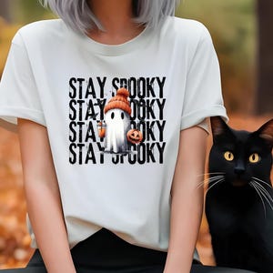 May include: White t-shirt with the text "STAY SPOOKY" stacked vertically. The graphic features a ghost wearing an orange beanie and holding a jack-o'-lantern and a lantern. A black cat with yellow eyes sits to the right.