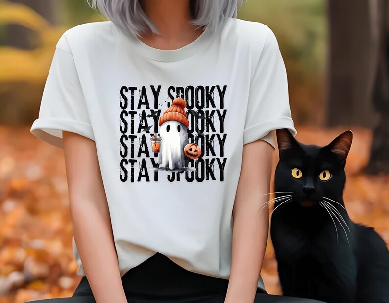 May include: White t-shirt with the text "STAY SPOOKY" stacked vertically. The graphic features a ghost wearing an orange beanie and holding a jack-o'-lantern and a lantern. A black cat with yellow eyes sits to the right.