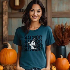 May include: Teal t-shirt with the text "Stay Spooky" stacked vertically. The shirt features a cartoon ghost wearing a witch hat and holding a pumpkin. Orange pumpkins are in the background, suggesting a Halloween theme.