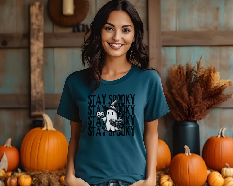 May include: Teal t-shirt with the text "Stay Spooky" stacked vertically. The shirt features a cartoon ghost wearing a witch hat and holding a pumpkin. Orange pumpkins are in the background, suggesting a Halloween theme.