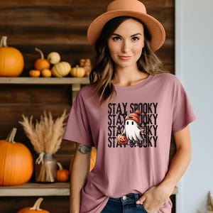May include: Mauve t-shirt with a Halloween-themed graphic. The graphic features a cartoon ghost wearing a knit cap and holding a pumpkin. The words "STAY SPOOKY" are stacked vertically in black text.