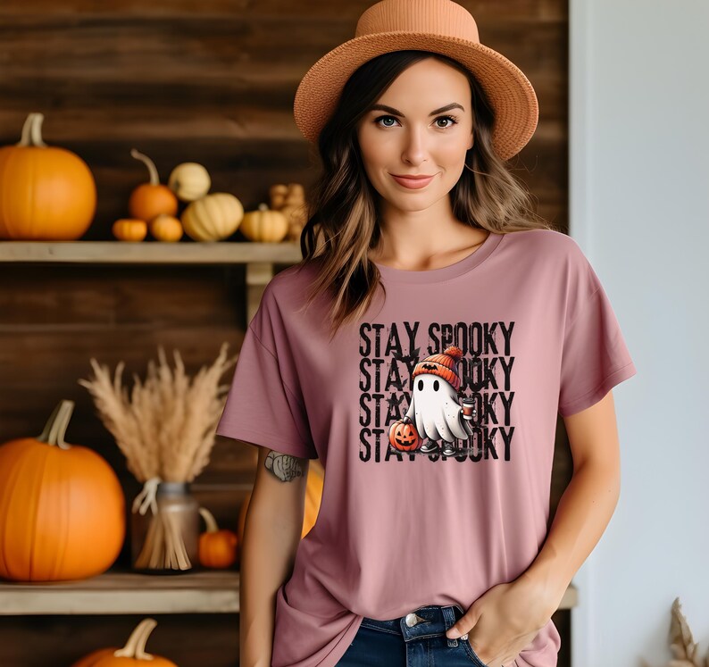 May include: Mauve t-shirt with a Halloween-themed graphic. The graphic features a cartoon ghost wearing a knit cap and holding a pumpkin. The words "STAY SPOOKY" are stacked vertically in black text.