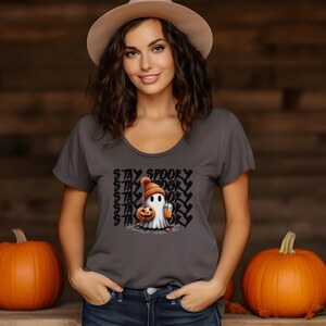 May include: A gray scoop-neck t-shirt featuring a cartoon ghost wearing an orange beanie and holding a pumpkin and a drink. The text "STAY SPOOKY" is printed above the ghost in a repeating pattern. Two orange pumpkins are in the background.