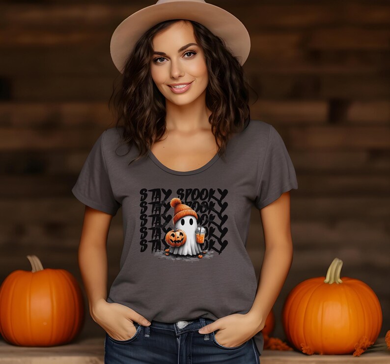 May include: A gray scoop-neck t-shirt featuring a cartoon ghost wearing an orange beanie and holding a pumpkin and a drink. The text "STAY SPOOKY" is printed above the ghost in a repeating pattern. Two orange pumpkins are in the background.