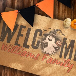 Puede incluir: Una alfombra de Halloween con la palabra "WELCOME" en letras marrones oscuras y "Williams Family" en naranja. Un fantasma de dibujos animados con un sombrero de bruja sostiene una calabaza. Guirnalda de banderines naranja y negro y calabazas completan la escena.