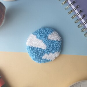 May include: A blue and white wool punch needle embroidery depicting a sky with clouds. The embroidery is circular and has a textured surface.