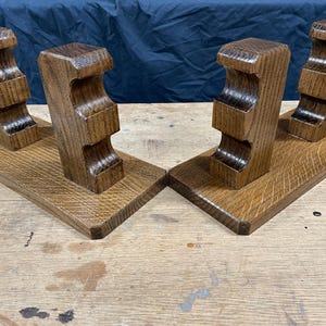 May include: Two matching wooden bookends. Each bookend has a rectangular base with a carved, decorative support. The wood has a warm, medium brown tone and a visible grain pattern. The bookends are designed to hold books upright.