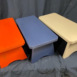 May include: Three wooden step stools painted in different colors: orange, blue, and cream. The stools are all the same size and shape, with a simple, rectangular top and a curved base.