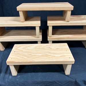 May include: A collection of wooden step stools in various sizes. The stools are made of light-colored wood and have a simple, functional design. The steps are rectangular with angled legs, and some have a shelf. The background is a dark blue fabric.