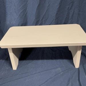 May include: A light beige wooden step stool with a simple design. The stool has a rectangular top and two short legs on each side.