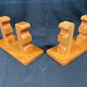 May include: Two wooden stands, each with a rectangular base and two upright supports. The supports have a curved design with two recessed areas. The wood has a light brown color.