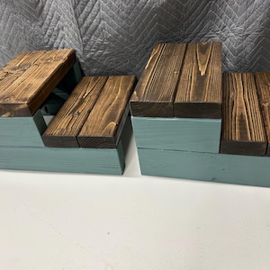 May include: Two wooden step stools. The steps are made of stained brown wood, and the base is painted a light blue-green. The stools are designed with a stepped appearance, with each step slightly smaller than the one below.
