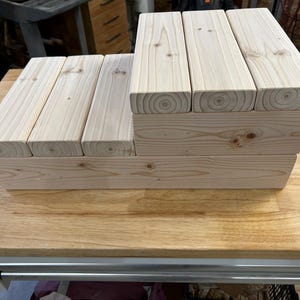 May include: A light-colored wooden tiered display stand featuring a stepped design with three lower platforms and three elevated platforms. The structure is constructed from natural pine wood planks showing visible grain patterns and circular knots.