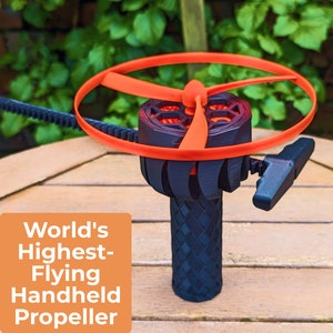 Op de afbeelding: Een zwart en oranje handbediend propeller speelgoed met een rode ring en een zwarte propeller. Het speelgoed ligt op een houten oppervlak. "World's Highest-Flying Handheld Propeller" staat op een oranje achtergrond.