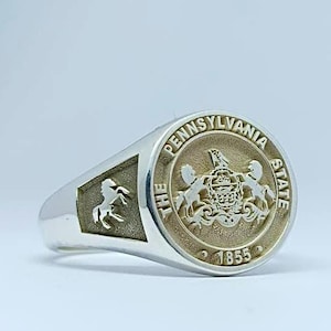 May include: A silver ring with a gold-toned seal featuring the Pennsylvania State University logo. The seal reads "The Pennsylvania State 1855".