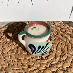 May include: A white ceramic candle holder with a green rim and blue and green floral designs. The candle holder is shaped like a pitcher and has a small handle. The candle holder is sitting on a woven straw mat.