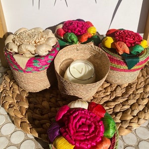 May include: Four woven baskets with colorful flower-shaped lids. The baskets are filled with candles and are sitting on a woven mat.