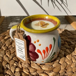 May include: A white ceramic mug with a yellow rim and colorful floral designs. The mug is filled with a white candle and a dried flower. The mug has a handle and a label that reads "Velas de Vela Candle Co. Vintage Gardenia 14oz Soy Candle".