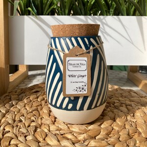 May include: A white ceramic candle jar with a blue and white striped design. The jar has a cork lid and a label that reads "Velas de Vela Candle Co. White Ginger 21 oz Soy Candle".