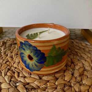 May include: A hand-painted terracotta candle holder with a floral design in shades of blue, green, and orange. The candle holder is filled with a white candle and has two wicks.