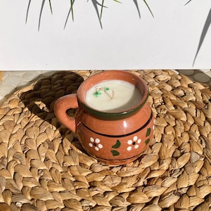 May include: A terracotta-colored mug candle with a white wax interior. The mug has a handle and is decorated with white flowers and green leaves. The rim is green. The candle sits on a woven, brown placemat.
