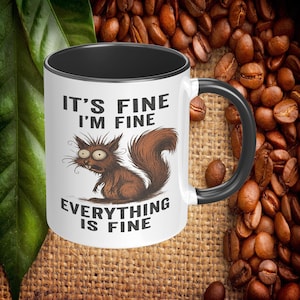 May include: White ceramic mug with a black interior and handle. The mug features a cartoon squirrel with the text "IT'S FINE I'M FINE EVERYTHING IS FINE". The squirrel is brown with wide eyes. The mug is surrounded by coffee beans.