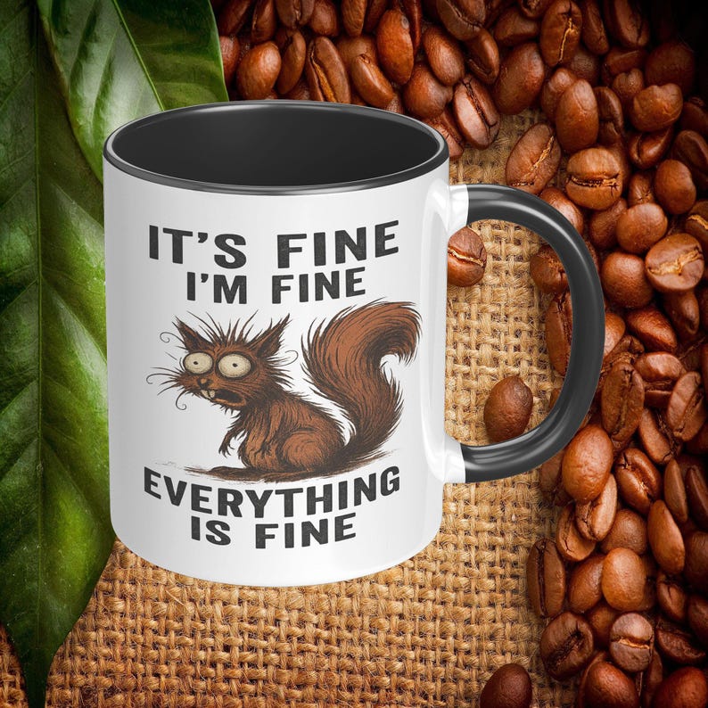 May include: White ceramic mug with a black interior and handle. The mug features a cartoon squirrel with the text "IT'S FINE I'M FINE EVERYTHING IS FINE". The squirrel is brown with wide eyes. The mug is surrounded by coffee beans.