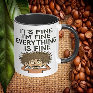 May include: White ceramic coffee mug with a black interior and handle. The mug features the text "IT'S FINE I'M FINE EVERYTHING IS FINE" with a cartoon illustration of a stressed person. The background includes coffee beans.