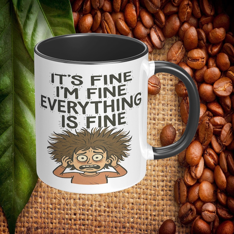 Pu&ograve; includere: Tazza da caff&egrave; in ceramica bianca con interno e manico neri. La tazza presenta la scritta "IT'S FINE I'M FINE EVERYTHING IS FINE" con un'illustrazione a cartoni animati di una persona stressata. Lo sfondo include chicchi di caff&egrave;.