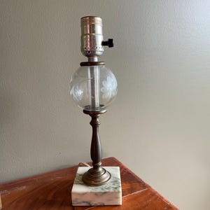 May include: A table lamp featuring a square white marble base with grey veining, a slender brass-toned metal stem, and a spherical clear glass globe etched with a floral pattern. The lamp includes a metallic socket with a black turn-knob switch.