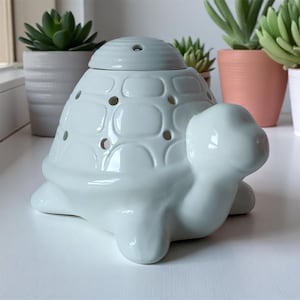 Peut inclure: Un chauffe-huile en céramique blanche en forme de tortue avec un couvercle amovible. La tortue a une carapace texturée avec plusieurs petits trous et une tête et des membres lisses et arrondis. Le chauffe-huile est conçu pour contenir des huiles parfumées.