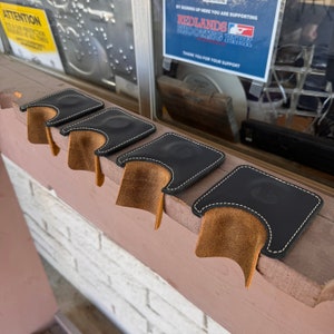 May include: Four black leather cartridge holders with white stitching and a brown interior. The holders are arranged in a row on a wooden surface. The image also shows a sign that says "ATTENTION BAR PROTECTION" and "REDLANDS SHOOTING PARK".