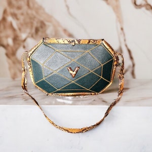 Könnte beinhalten: Eine geometrische Handtasche in Grün und Gold mit einem Schlangenlederriemen. Die Tasche hat eine Diamantenform und ein goldenes V in der Mitte.