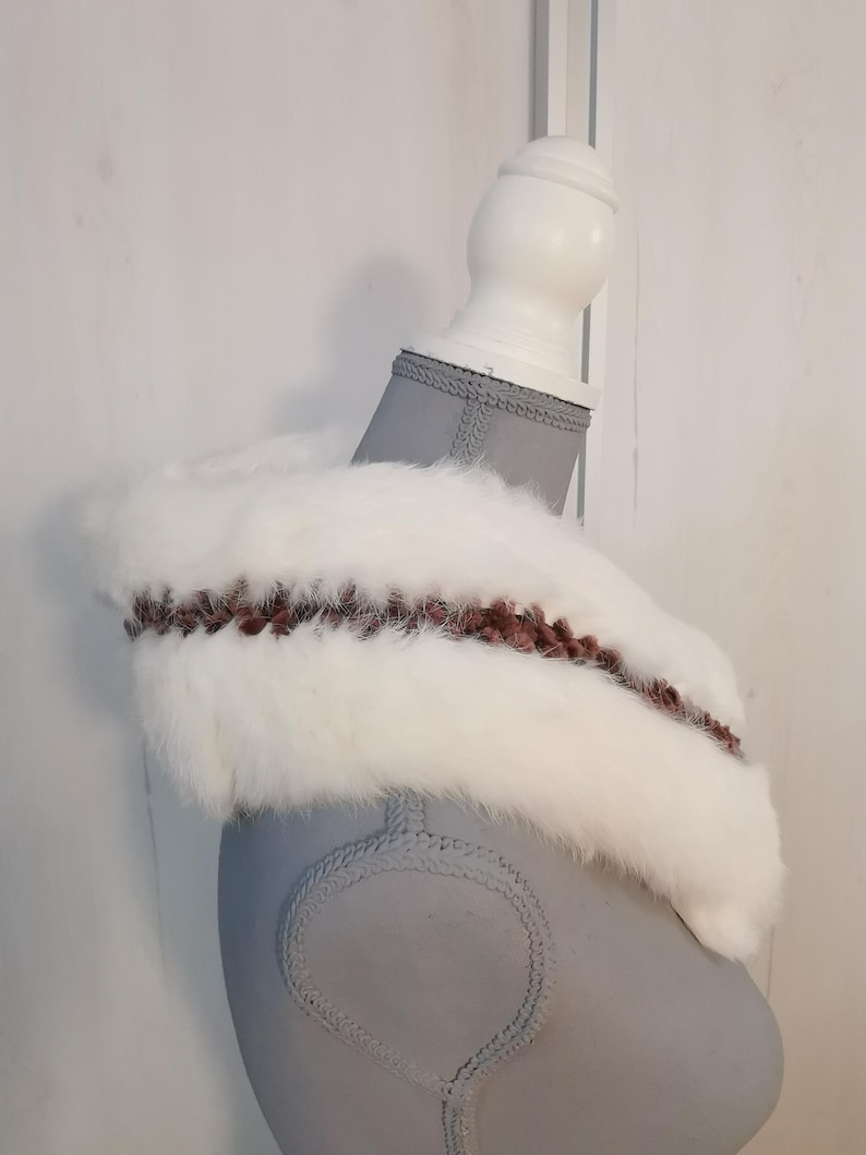 May include: White faux fur collar with a brown and pink braided trim. The collar is displayed on a gray mannequin.