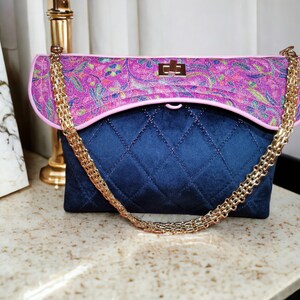 Puede incluir: Un bolso acolchado azul con una solapa de estampado floral rosa y una correa de cadena dorada.