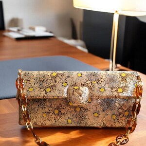 Cartera bolso exclusivo de piel genuina de tafilete, estampado floral holográfico, cierre broche forrado y cadena dorada y resina ámbar.