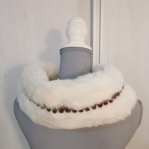 May include: White fur collar with a brown beaded trim. The collar is displayed on a gray mannequin.