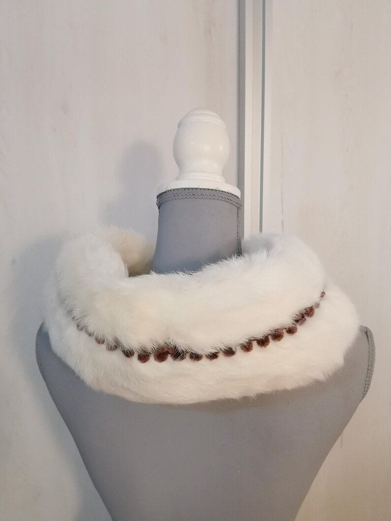 May include: White fur collar with a brown beaded trim. The collar is displayed on a gray mannequin.