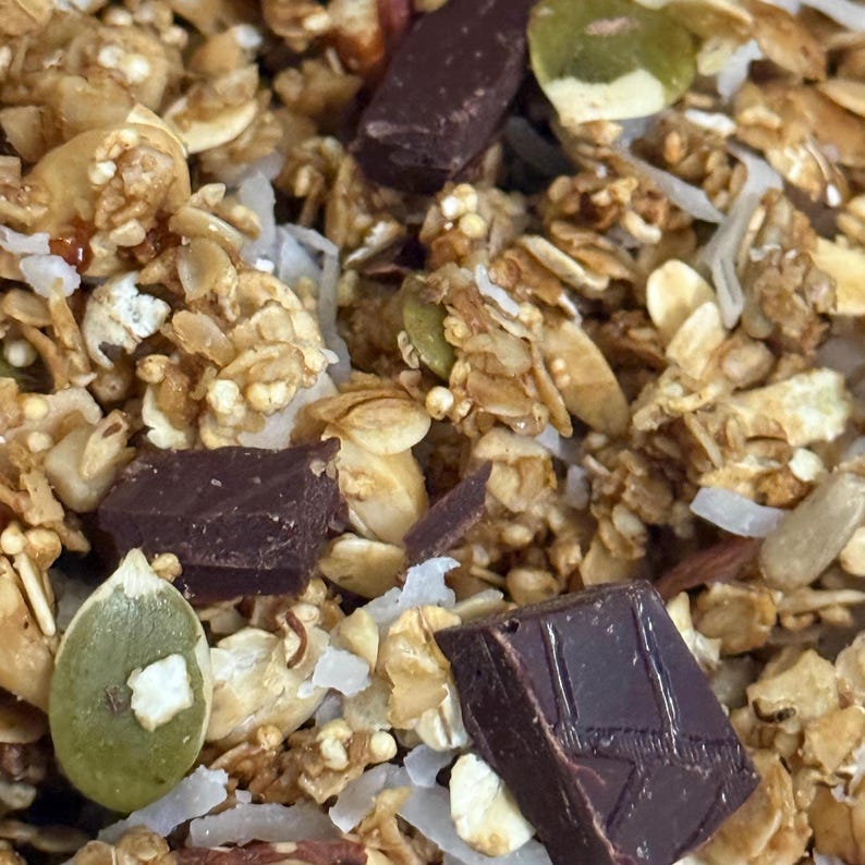 May include: A close-up shot of granola with dark chocolate pieces, pumpkin seeds, and coconut flakes. The granola is golden brown and crunchy, with varied textures and colours. The chocolate is dark brown, and the seeds are green.