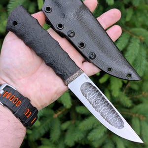 以下が含まれることがあります： 黒いテクスチャ加工のハンドルと、暗い斑点模様とN690の文字が入った銀色の刃を持つ固定刃ナイフ。ナイフには、端に複数の円形のハトメが付いた、お揃いの黒いテクスチャ加工のシースが含まれています。