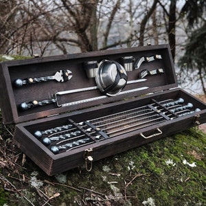 May include: A wooden box with a hinged lid, containing a set of metal skewers and grilling tools. The box is open, revealing the skewers and tools inside. The tools include a spatula, a grill brush, and a set of skewers.