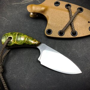 Può includere: Un piccolo coltello con manico verde e marrone e lama argentata. Il coltello è attaccato a un cordino marrone e si trova su una superficie grigio scuro. Nell'immagine è presente anche un fodero in pelle marrone con occhielli neri e un cordino marrone.