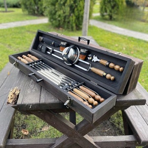 May include: A wooden carrying case with a black finish, containing a set of metal skewers with wooden handles, a metal spatula, and a small metal bowl. The case is open and the skewers are arranged neatly inside.