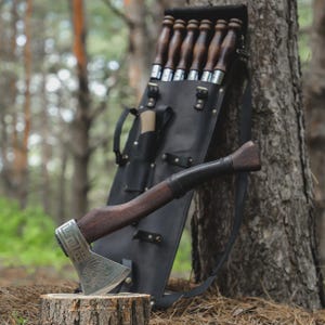 May include: Axe with a wooden handle and a silver blade featuring an engraved design. A black leather sheath holds several skewers and a small knife. The axe rests on a tree stump, with the set leaning against a tree in a forest setting.