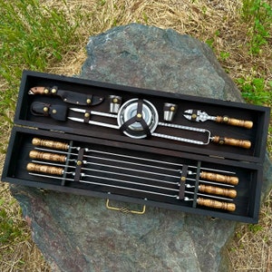 May include: A wooden box with a black interior containing a set of grilling tools. The tools include skewers, a spatula, a knife, and a small metal cup. The box is open and the tools are arranged neatly inside.