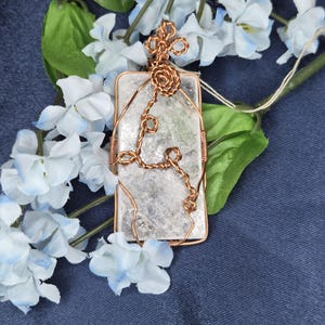 May include: A rectangular clear crystal pendant encased in a decorative copper wire wrap. The wire features intricate coiled details and a looped bail at the top for suspension. The crystal has a textured, semi-translucent appearance with visible internal inclusions.