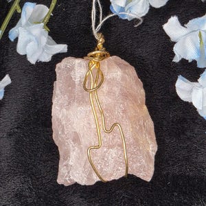 May include: A raw, light pink rose quartz pendant with a gold-colored wire wrap. The pendant is suspended by a thin, off-white string. The background is black with light blue flowers.
