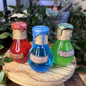 Può includere: Tre bottiglie di pozione con liquidi rossi, blu e verdi. Ogni bottiglia ha un sigillo di cera e un'etichetta con la scritta "Health+", "Mana" e "Stamina". Le bottiglie sono su un vassoio di legno, con uno sfondo di fogliame verde.